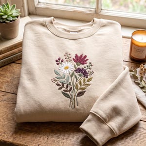 Op de afbeelding: Een beige sweater met ronde hals en een centraal geborduurd boeket wilde bloemen. Het ontwerp bevat een witte margriet met een geel hart, een donkerrood blad, paarse bessen en diverse groene bladeren en stengels in een verticaal boeket.