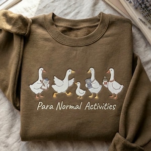 Puede incluir: Sudadera marrón con un diseño bordado de cinco gansos blancos. Los gansos están representados en diferentes poses, uno con un portapapeles y otro con una taza de café. El texto "Para Normal Activities" está debajo.