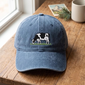 Puede incluir: Una gorra de béisbol azul con un gráfico bordado de una vaca blanca y negra pastando sobre un trozo de hierba verde. La gorra tiene una corona estructurada, una visera curva y ojales cosidos en los paneles superiores para ventilación.