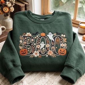 May include: A dark green crewneck sweatshirt featuring an embroidered design of the word BOO in large letters. The letters are decorated with orange pumpkins, white ghosts, black bats, a spider web, and various floral elements in autumn colors.
