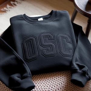 May include: A black sweatshirt with the letters "OSC" in a raised, textured design across the chest. The sweatshirt has a crew neck and long sleeves with ribbed cuffs. The garment is folded on a woven surface.