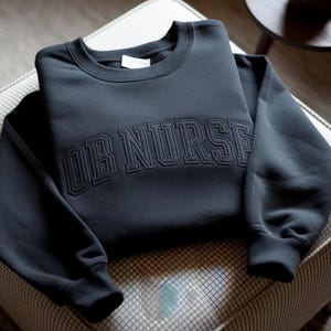 Op de afbeelding: Een donkergrijs sweatshirt met de geborduurde tekst "OB NURSE" over de borst. De crewneck sweatshirt is op een patroon oppervlak gevouwen. De mouwen zijn aan de polsen geribbeld.