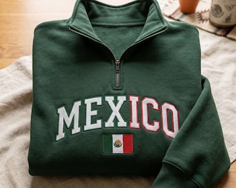 May include: A dark green quarter-zip sweatshirt featuring the word MEXICO embroidered in a white and red arched font across the chest. Below the text is a small, rectangular embroidered patch of the Mexican flag with its green, white, and red stripes.