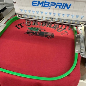 May include: An industrial embroidery machine with the brand name EMBPRIN is stitching a design onto red fabric held in a green hoop. The design features a black truck carrying a green pine tree with the text IT'S A BEAUT in black lettering.