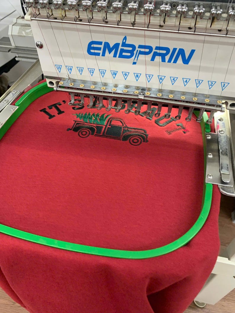 May include: An industrial embroidery machine with the brand name EMBPRIN is stitching a design onto red fabric held in a green hoop. The design features a black truck carrying a green pine tree with the text IT'S A BEAUT in black lettering.
