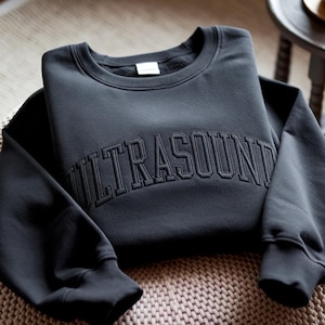 Puede incluir: Una sudadera gris oscuro con la palabra "ULTRASOUND" bordada en el pecho en un tono a juego. Tiene cuello redondo y puños y dobladillo acanalados. La sudadera está doblada sobre una superficie texturizada.