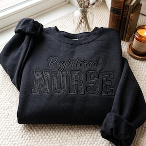 May include: A black crewneck sweatshirt featuring the embroidered text Registered NURSE. The word Registered is in a cursive script font, while the word NURSE is in large block letters filled with a detailed, tonal floral and botanical pattern.