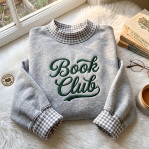 May include: A heather grey sweatshirt featuring a mock neck collar and sleeve cuffs in a white and grey gingham pattern. The front displays the text "Book Club" embroidered in dark green cursive lettering with a bold, rounded font style.