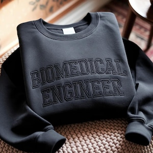 Könnte beinhalten: Ein dunkelgraues Sweatshirt mit dem gestickten Schriftzug "BIOMEDICAL ENGINEER" in passendem Dunkelgrau auf der Brust. Das Sweatshirt ist ordentlich auf einer gewebten Oberfläche gefaltet, wobei die Ärmel und der Halsausschnitt sichtbar sind.