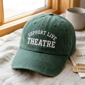 May include: A forest green baseball cap featuring the white embroidered text SUPPORT LIVE THEATRE in a curved arch across the front panels. The cap has a structured crown, a curved brim, and stitched eyelets on the top panels for ventilation.