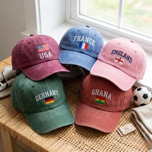 Op de afbeelding: Vijf baseballpetten in verschillende kleuren, elk voorzien van een geborduurde landnaam en de bijbehorende nationale vlag. De petten zijn bordeauxrood, blauw, roze, groen en terracotta, met de teksten USA, FRANCE, ENGLAND, GERMANY en GHANA.