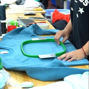 May include: A teal sweatshirt secured in a green embroidery hoop on a wooden table. A second green embroidery hoop rests on a light blue fabric in the foreground. The sweatshirt features a crew neckline and long sleeves, prepared for machine embroidery.