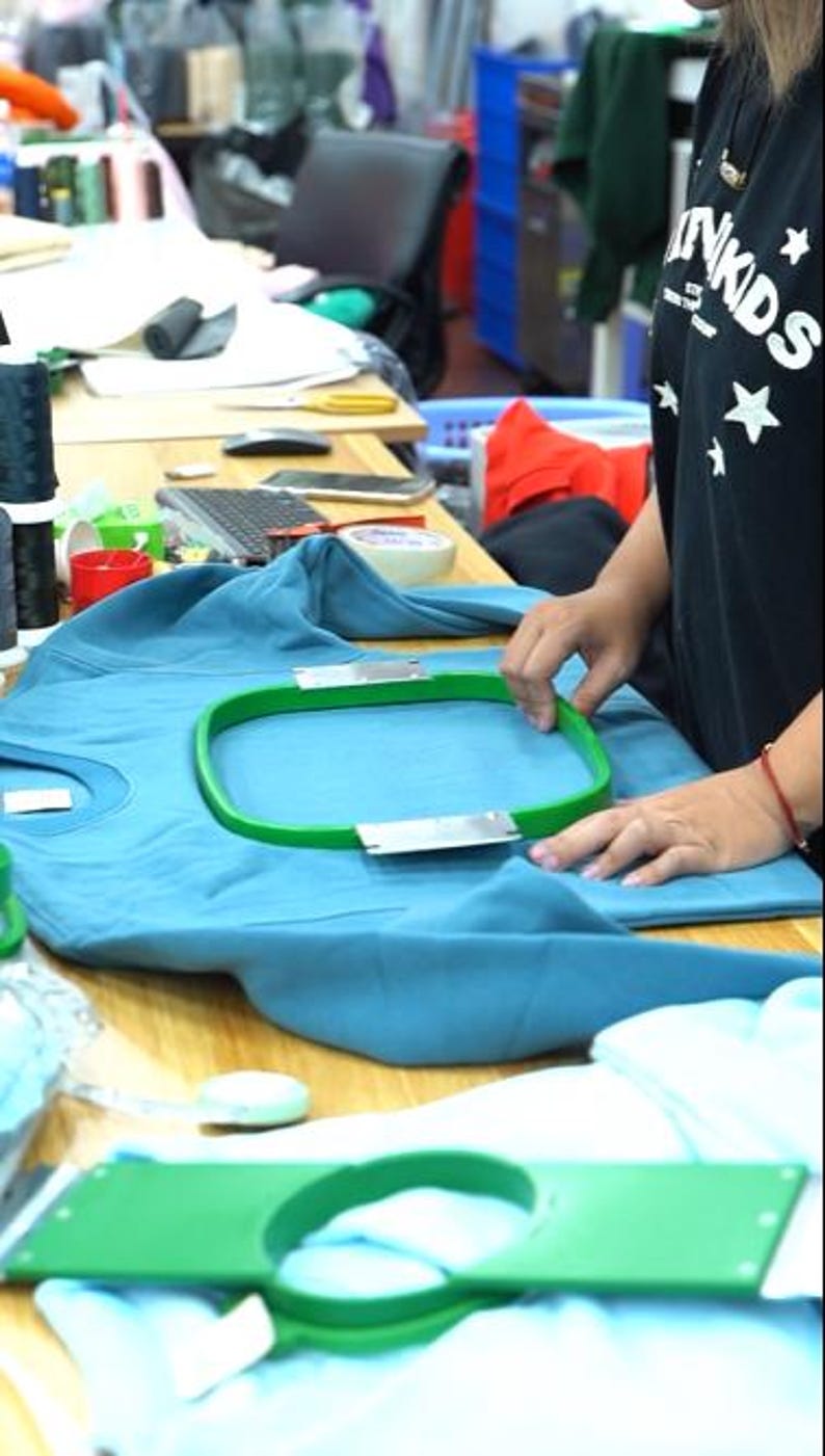 May include: A teal sweatshirt secured in a green embroidery hoop on a wooden table. A second green embroidery hoop rests on a light blue fabric in the foreground. The sweatshirt features a crew neckline and long sleeves, prepared for machine embroidery.