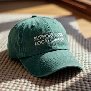 Peut inclure: Casquette de baseball verte avec le texte brodé "SUPPORT YOUR LOCAL LIBRARY" en blanc. La casquette est faite d'un tissu doux et a une visière incurvée. Le chapeau est posé sur une surface texturée.