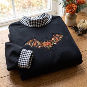 May include: A black long-sleeve sweatshirt featuring a mock neck collar and sleeve cuffs with a black and white gingham pattern. The center of the chest displays an embroidered bat silhouette filled with intricate floral details in orange, red, and green.