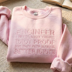 Może przedstawiać: Jasnoróżowa bluza z wyhaftowanym słowem "ENGINEER" oraz napisem: "I TRY TO MAKE THINGS IDIOT PROOF BUT THEY KEEP MAKING BETTER IDIOTS."