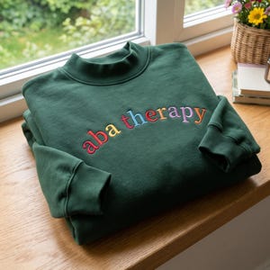 May include: A dark forest green long-sleeve sweatshirt featuring a mock neck collar and ribbed cuffs. The chest displays the text "aba therapy" embroidered in a curved arc, with each individual letter stitched in a different vibrant color.