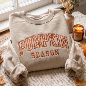 May include: A beige crewneck sweatshirt featuring the text PUMPKIN SEASON embroidered across the chest. The word PUMPKIN is filled with a patterned fabric featuring small orange and brown pumpkins, while the word SEASON is in solid orange thread.