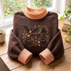 May include: A brown long-sleeve sweatshirt featuring a mock neck collar and sleeve cuffs in an orange and white gingham pattern. The center of the chest displays an embroidered design of four black bats, a crescent moon, and various flowers.