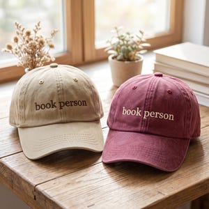 May include: Two baseball caps, one tan and one maroon, each featuring the text "book person" embroidered on the front. The tan cap has dark brown lettering, while the maroon cap has light cream-colored lettering. Both caps have a curved brim.