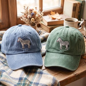 May include: Two baseball caps, one in a faded blue and one in a muted green, each featuring an embroidered zebra graphic on the front panel. Both caps have a structured crown, a curved brim with stitched detailing, and eyelets for ventilation.