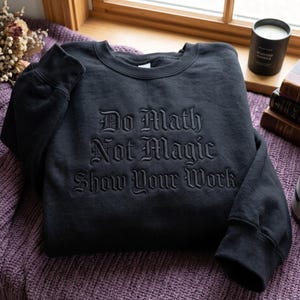 May include: A black crewneck sweatshirt featuring black tonal embroidery on the chest. The text reads "Do Math Not Magic Show Your Work" in a gothic-style font. The garment has long sleeves with ribbed cuffs and a ribbed neckline.