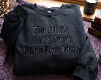 May include: A black crewneck sweatshirt featuring black tonal embroidery on the chest. The text reads "Do Math Not Magic Show Your Work" in a gothic-style font. The garment has long sleeves with ribbed cuffs and a ribbed neckline.