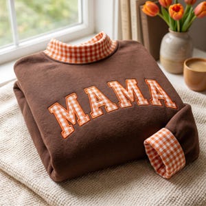 May include: A brown sweatshirt featuring the word MAMA in orange and white gingham block letters. The garment has a matching orange and white gingham mock neck collar and folded sleeve cuffs, showcasing a consistent plaid pattern.