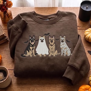 May include: A brown crewneck sweatshirt featuring a graphic print of four German Shepherd dogs. One dog is dressed as a ghost, one as a mummy, and two are sitting normally. Small black bats are scattered around the dogs on the front of the garment.