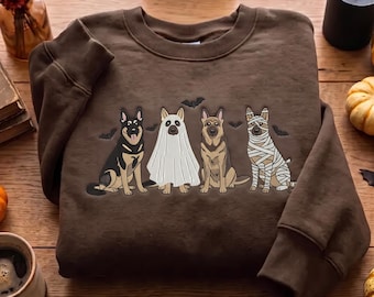 May include: A brown crewneck sweatshirt featuring a graphic print of four German Shepherd dogs. One dog is dressed as a ghost, one as a mummy, and two are sitting normally. Small black bats are scattered around the dogs on the front of the garment.