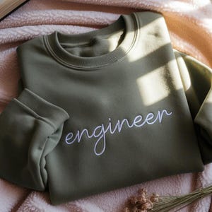 以下が含まれることがあります： 白い筆記体で「engineer」と刺繍されたオリーブグリーンのスウェットシャツ。スウェットシャツは折りたたまれており、袖と襟が見えます。生地は柔らかく快適に見えます。