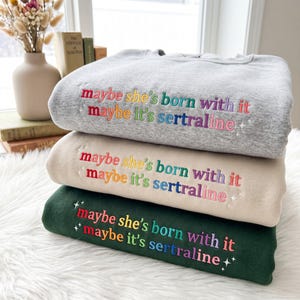 Może przedstawiać: Trzy bluzy z okrągłym dekoltem w kolorze jasnoszarym, beżowym i ciemnozielonym. Każda posiada wyhaftowany tęczowy napis "maybe she's born with it maybe it's sertraline" oraz małe wyhaftowane gwiazdki otaczające tekst.
