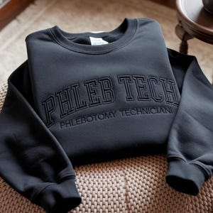 May include: A dark grey sweatshirt featuring the embroidered words "PHLEB TECH" above "PHLEBOTOMY TECHNICIAN". The sweatshirt is folded on a woven surface. The sleeves are visible on either side of the folded garment.