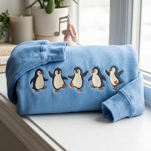 Könnte beinhalten: Ein hellblaues Sweatshirt mit einer Stickerei von fünf einzelnen Pinguinen in verschiedenen Tanzposen. Jeder Pinguin ist mit einem schwarz-weißen Körper und orangefarbenen Füßen dargestellt, die in einer horizontalen Reihe auf der Vorderseite angeordnet sind.
