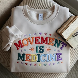 May include: Cream-colored sweatshirt with the embroidered text "MOVEMENT IS MEDICINE" in rainbow colors. The design includes hearts and stars. A book and lavender sprig are visible in the background.