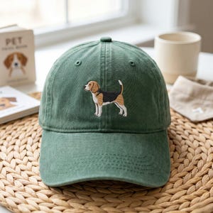 Op de afbeelding: Een bosgroene baseballpet met een geborduurde beagle op het voorpaneel. De hond is in profiel afgebeeld, staand met witte, bruine en zwarte vachtkleuren. De pet heeft een gebogen klep, een knoop aan de bovenkant en gestikte oogjes.