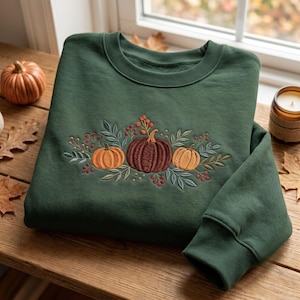 May include: A forest green crewneck sweatshirt featuring an embroidered design of three pumpkins in shades of orange and brown, surrounded by detailed green leaves and small red berries arranged in a horizontal cluster across the chest.