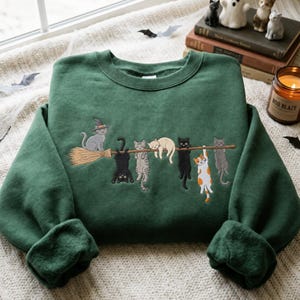 Op de afbeelding: Een donkergroene sweater met ronde hals en een borduursel van zeven katten die aan een houten bezemsteel hangen. Eén kat draagt een heksenhoed, terwijl de andere katten zijn afgebeeld in kleuren zoals zwart, grijs, crème en oranje-wit.