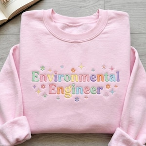 以下が含まれることがあります： 「Environmental Engineer」の文字がカラフルな刺繍で施された、薄いピンク色のスウェットシャツ。小さな花と星のアクセントがデザインされています。柔らかい素材で作られたスウェットシャツです。