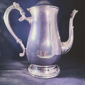 May include: A silver coffee pot with a curved handle and spout. The pot has a decorative base and lid, set against a dark background. The pot is likely made of metal and has a reflective surface. The pot is an antique.