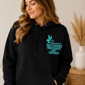 Op de afbeelding: Een zwarte hoodie met een turquoise afbeelding op de linkerborst. Het ontwerp bevat het silhouet van een fee en de tekst: Powered by BITCHDUST CUSS WORDS AND CAFFEINE. De trui is voorzien van een kangoeroezak en zwarte trekkoorden.