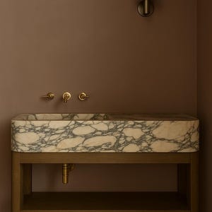 May include: A rectangular marble sink with gray veining sits on a wooden stand with a lower shelf. The sink has brass fixtures and is mounted against a brown wall. A brass sconce is mounted on the wall above the sink.
