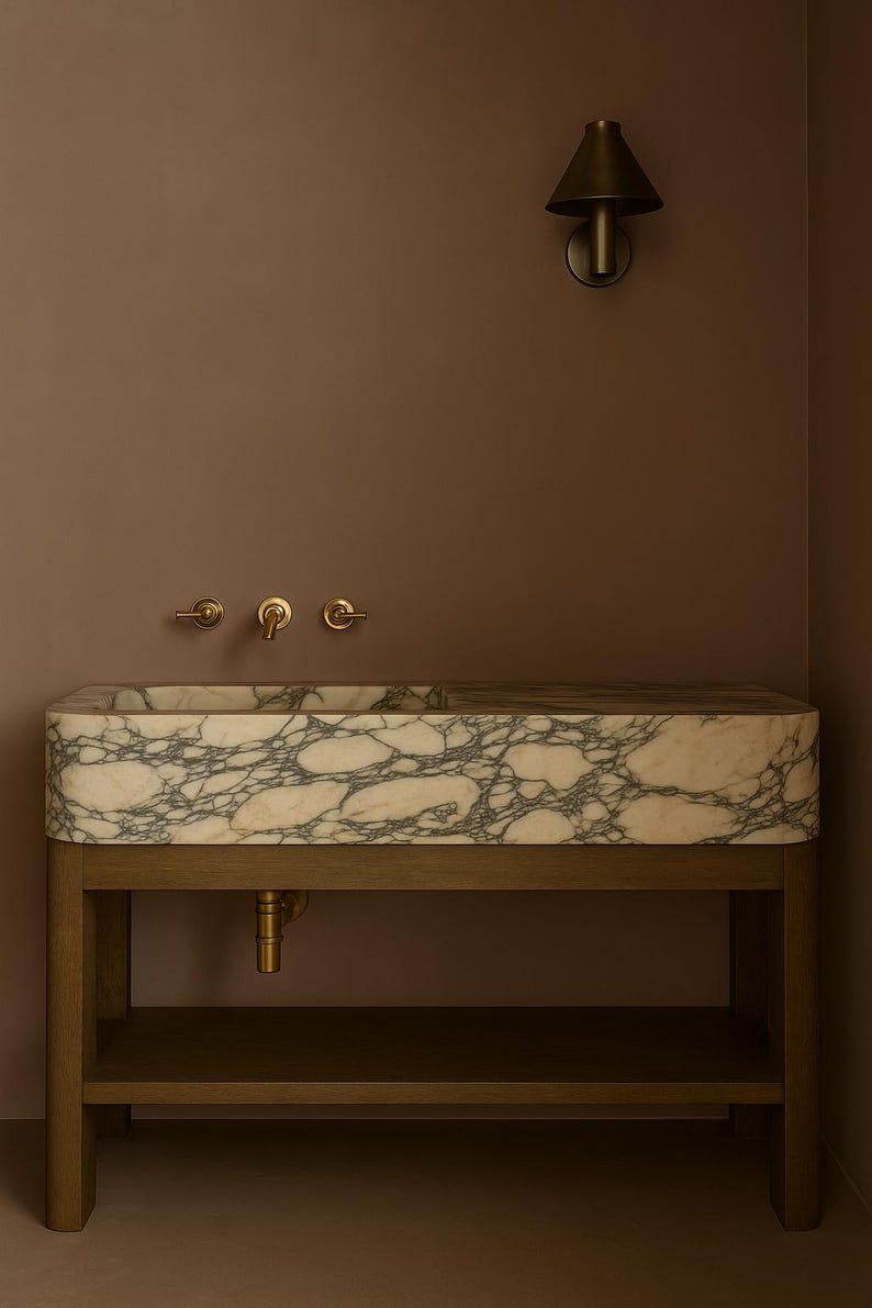 May include: A rectangular marble sink with gray veining sits on a wooden stand with a lower shelf. The sink has brass fixtures and is mounted against a brown wall. A brass sconce is mounted on the wall above the sink.