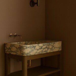 May include: A marble sink with a light brown and gray pattern sits on a wooden stand with a lower shelf. The sink has a brass faucet and is mounted against a brown wall. A matching wall sconce is above the sink.
