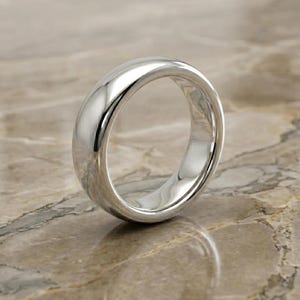 Könnte beinhalten: Ein polierter Silberring mit einem abgerundeten Band. Der Ring ist hochreflektierend und zeigt die Umgebung. Der Ring ist vor einem neutralen Hintergrund platziert.