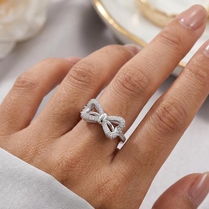 May include: A silver-colored ring in the shape of a bow, encrusted with small, clear gemstones. The ring is on a person's finger, with the hand in the foreground. The background is blurred, with a hint of a white flower and a gold-rimmed dish.