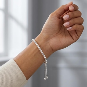 May include: A silver beaded bracelet with a sliding clasp is worn on a wrist. The bracelet features small, round beads and a delicate chain with two small ball charms at the end. The wrist is adorned with a light-colored sweater.