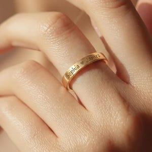 Op de afbeelding: Een goudkleurige bandring met een gladde, gepolijste afwerking. De buitenkant van de ring is gegraveerd met de namen Sophia en James, gescheiden door een klein hartsymbool. De ring heeft een eenvoudige, gelijkmatige breedte en een rond profiel.