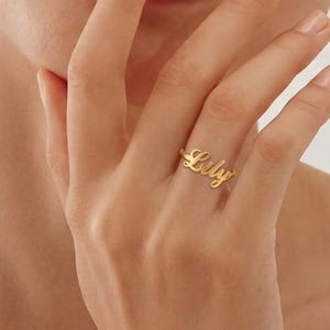 Op de afbeelding: Een goudkleurige ring met de naam Lily geschreven in een cursief lettertype. De ring heeft een gepolijste afwerking en wordt gedragen aan de ringvinger, waarbij het fijne metaalwerk van de letters die verbonden zijn met de dunne ringband zichtbaar is.