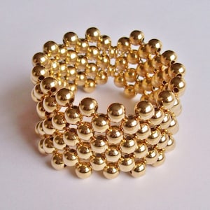 Op de afbeelding: Een gouden armband gemaakt van kleine, ronde kralen. De kralen zijn gerangschikt in een patroon dat een textuur creëert.